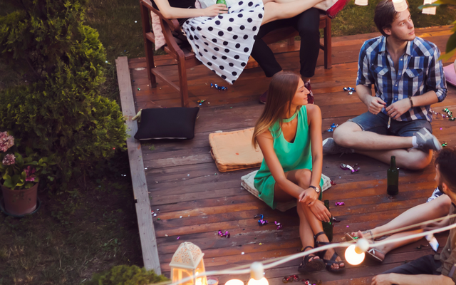 Eine Gruppe junger Erwachsener sitzt abends entspannt auf einer Holzterrasse im Freien, beleuchtet von Lichterketten und Laternen. Einige sitzen auf einer Bank, andere auf Kissen auf dem Boden. Konfetti liegt verstreut herum.