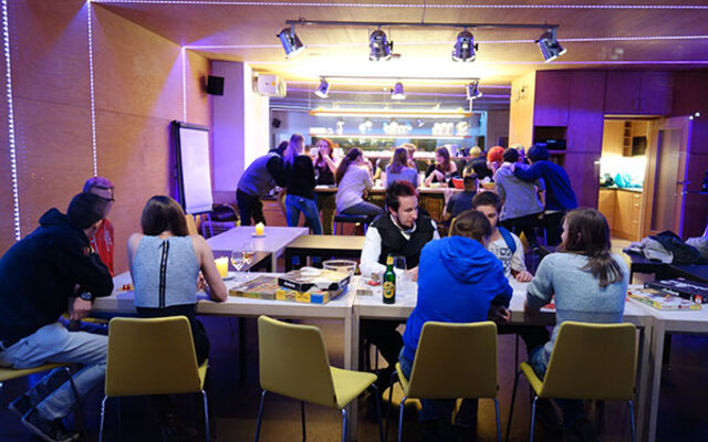 Das Bild zeigt eine Gruppe junger Menschen, die in einer gemütlichen und modernen Umgebung zusammenkommen. Im Vordergrund sitzen mehrere Personen an Tischen und spielen Brettspiele oder unterhalten sich. Im Hintergrund versammeln sich weitere Menschen an einer Bar oder Theke, was auf eine gemeinschaftliche Veranstaltung oder ein Treffen hindeutet. Die Beleuchtung mit LED-Streifen und Spots verleiht dem Raum eine warme, freundliche Atmosphäre.